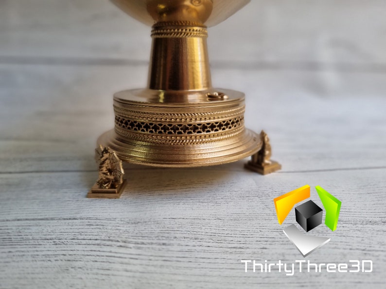 May include: A gold-colored metal decorative stand with a round top and three legs. The stand has a detailed design with a patterned base and a decorative top.