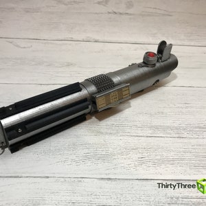 May include: A silver lightsaber prop with a black grip and a red button. The lightsaber has a gold detail on the side with a series of numbers.