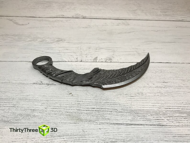 May include: A silver-colored, curved, metal knife with a decorative pattern on the blade. The knife has a ring at the top of the handle.