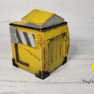 May include: A small, yellow, weathered, cube-shaped box with a black top and a black and white striped panel on the side. The box has a slightly open top and appears to be made of plastic.