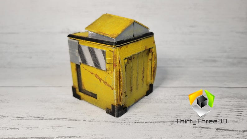 May include: A small, yellow, weathered, cube-shaped box with a black top and a black and white striped panel on the side. The box has a slightly open top and appears to be made of plastic.
