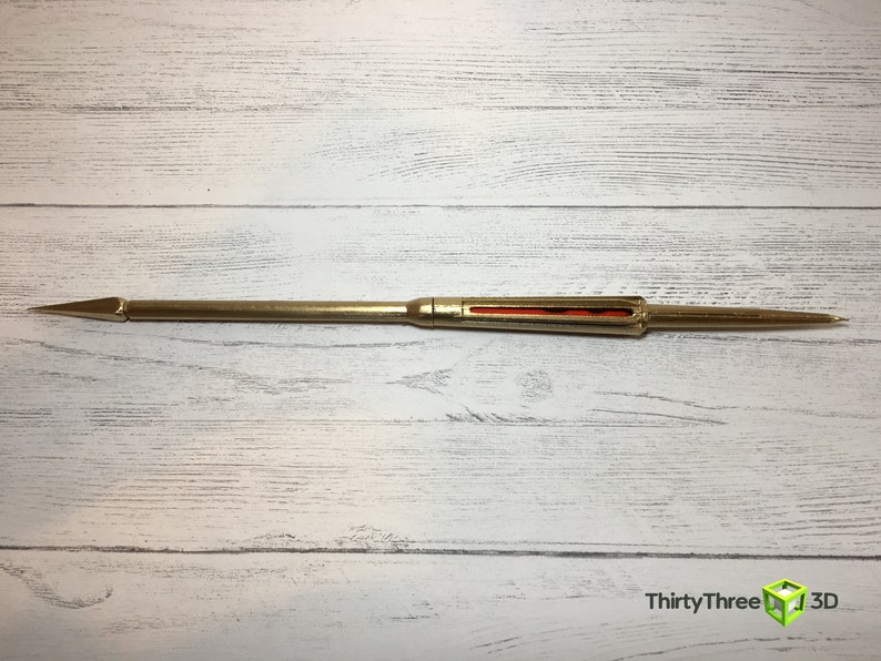 May include: A gold-colored metal dart with a red and orange stripe in the middle. The dart is sharp at one end and has a rounded tip at the other end.