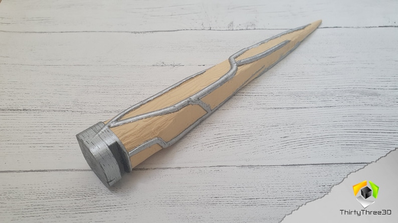 Op de afbeelding: Een zilveren en beige 3D-geprint rekwisietwapen met een puntige punt en een getextureerd oppervlak.