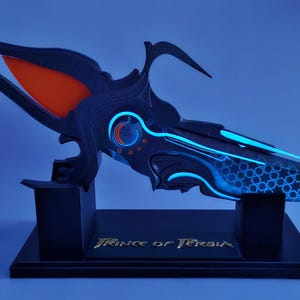 May include: A futuristic dagger prop in black and blue, with an orange accent. The dagger is displayed on a black stand with the text "Prince of Persia" in gold. The blade has a honeycomb pattern. The overall design is intricate and detailed.