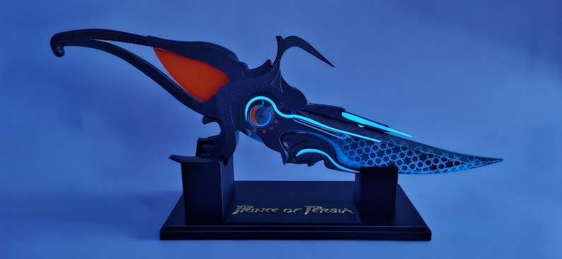 May include: A futuristic dagger prop in black and blue, with an orange accent. The dagger is displayed on a black stand with the text "Prince of Persia" in gold. The blade has a honeycomb pattern. The overall design is intricate and detailed.