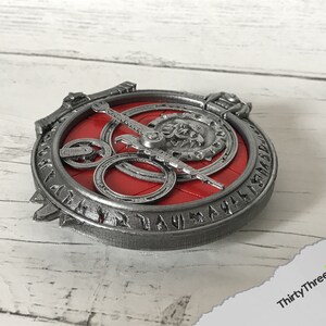May include: A silver-coloured metal pendant with a red centre and intricate details. The pendant features a circular design with a central symbol and surrounding runes.
