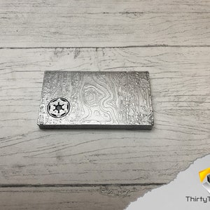 May include: A silver metal plate with a raised, textured design. The plate features a circular cutout in the center with a star-shaped symbol inside.