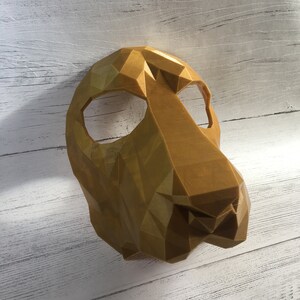 May include: A gold-colored, geometric, 3D printed animal mask. The mask has a triangular shape and is designed to cover the eyes and nose.