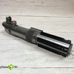 May include: A silver lightsaber prop with a black handle and a red button on the top. The lightsaber is on a white wooden surface.