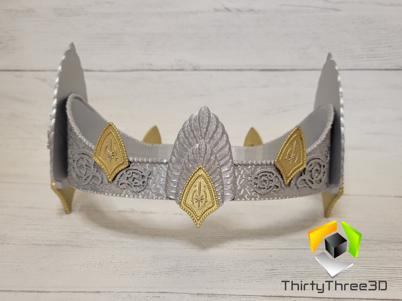 May include: A silver crown with gold accents and a detailed design. The crown has a pointed top and a band with intricate patterns.