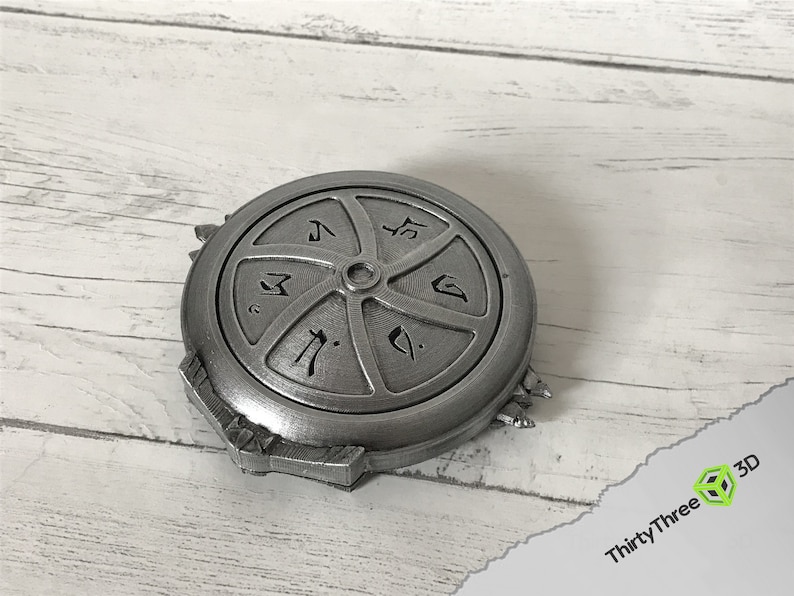 May include: A silver-colored metal disc with a circular design and symbols. The disc has a raised center and a textured surface.