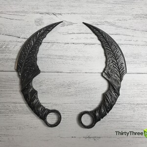 Puede incluir: Un par de cuchillos de lanzamiento de metal negro con dise&ntilde;os intrincados. Los cuchillos son curvos y tienen un anillo al final del mango.