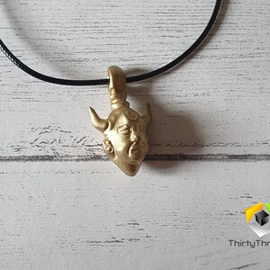 May include: A gold-colored pendant necklace featuring a detailed, three-dimensional sculpture of a horned, human-like face. The pendant is hanging from a black cord.