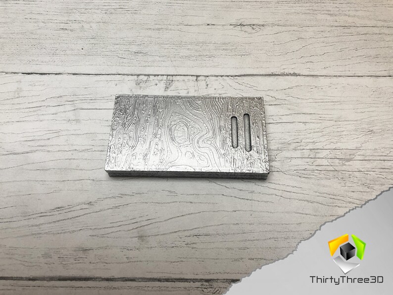 May include: A silver metal plate with a wood grain pattern and two rectangular cutouts. The plate is on a white wooden surface.