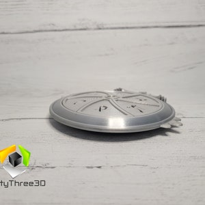 Puede incluir: Un objeto redondo de color plateado, impreso en 3D, con un dise&ntilde;o circular y un borde elevado. El objeto tiene una superficie texturizada y parece ser una pieza decorativa.