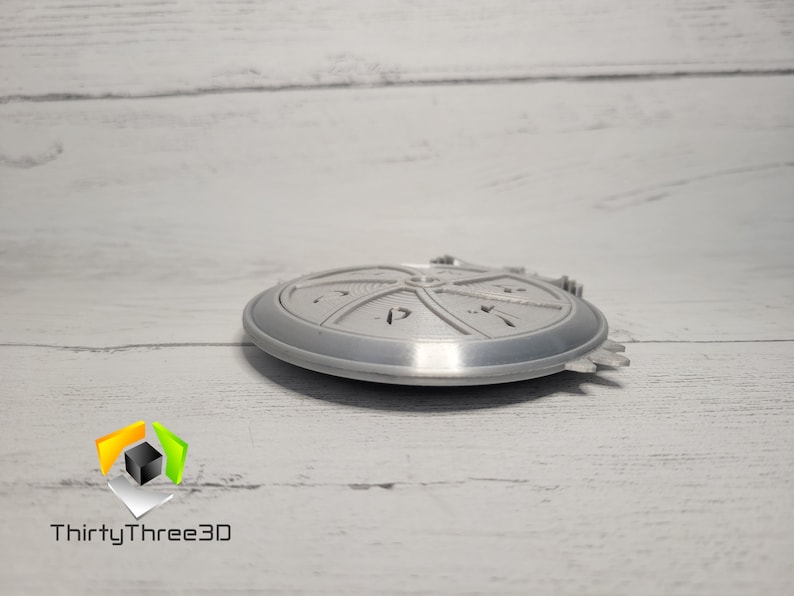 Puede incluir: Un objeto redondo de color plateado, impreso en 3D, con un dise&ntilde;o circular y un borde elevado. El objeto tiene una superficie texturizada y parece ser una pieza decorativa.