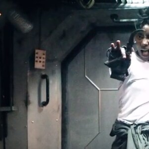 May include: A person in a white shirt and gray pants, holding a futuristic gun and gesturing with their other hand. The scene is set in a dimly lit, industrial-looking interior with another person in a black hooded garment.