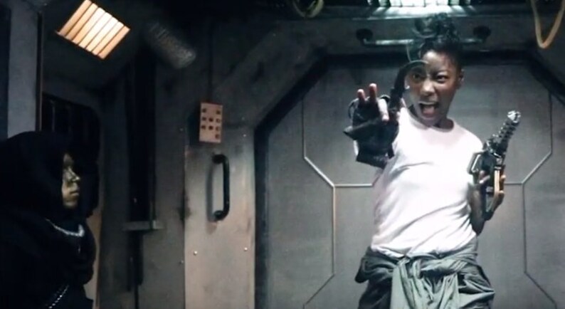May include: A person in a white shirt and gray pants, holding a futuristic gun and gesturing with their other hand. The scene is set in a dimly lit, industrial-looking interior with another person in a black hooded garment.
