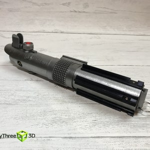 May include: A silver lightsaber prop with a black handle and a red button on the top. The lightsaber is on a white wooden surface.