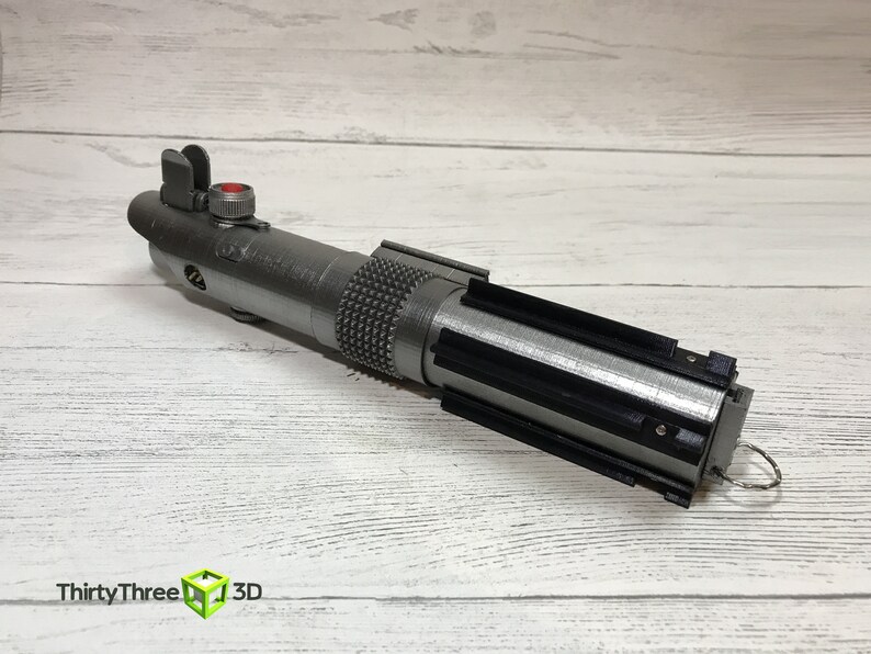 May include: A silver lightsaber prop with a black handle and a red button on the top. The lightsaber is on a white wooden surface.