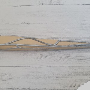 Op de afbeelding: Een zilveren en beige 3D-geprint rekwisiet dat lijkt op een skeletachtige arm, met een puntig uiteinde.