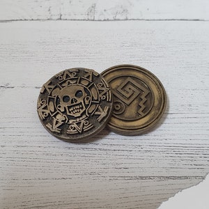 May include: Two gold-colored metal coins with intricate designs. One coin features a skull and crossbones, while the other has a geometric pattern.