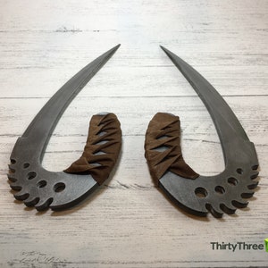 May include: Two silver-colored, curved, metal blades with brown leather handles. The blades have a serrated edge and multiple holes. The handles are wrapped in brown leather with a criss-cross pattern. The blades are displayed on a white wooden surface.