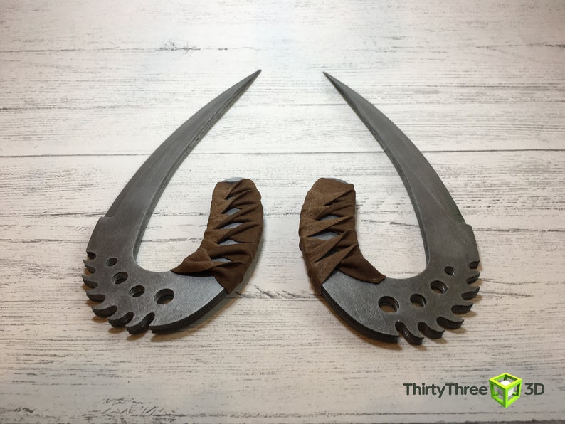 May include: Two silver-colored, curved, metal blades with brown leather handles. The blades have a serrated edge and multiple holes. The handles are wrapped in brown leather with a criss-cross pattern. The blades are displayed on a white wooden surface.