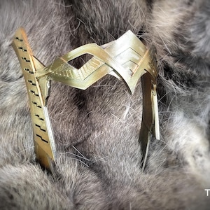 May include: A gold-colored headband with a geometric design. The headband is made of metal and has a smooth finish.