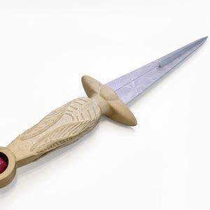 May include: A decorative dagger with a light-colored wooden handle and a silver blade. The handle features carved details and a red gemstone set in the pommel. The blade has a patterned design.