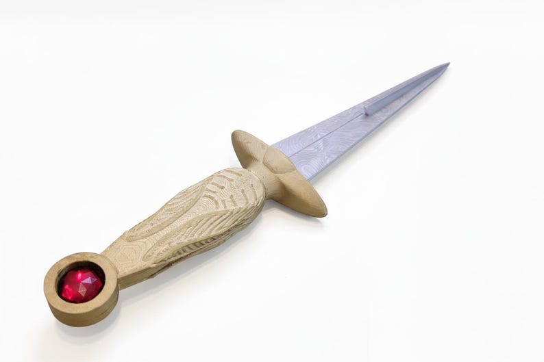 May include: A decorative dagger with a light-colored wooden handle and a silver blade. The handle features carved details and a red gemstone set in the pommel. The blade has a patterned design.