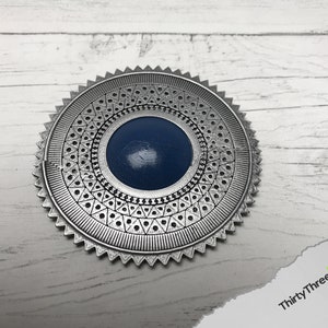 May include: A silver-colored metal medallion with a blue center and intricate geometric designs around the edge.