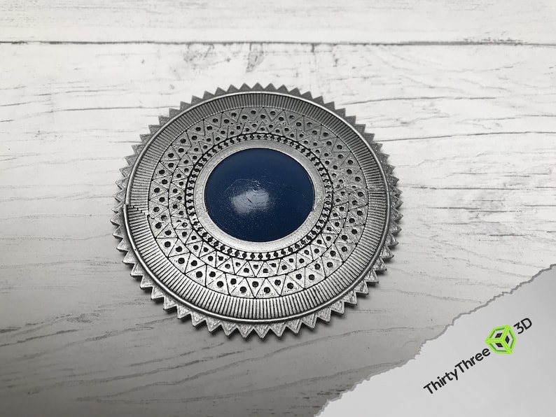 May include: A silver-colored metal medallion with a blue center and intricate geometric designs around the edge.