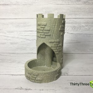 May include: A gray stone tower-shaped dice rolling tower with a brick pattern. The tower has a small opening at the top and a larger opening at the bottom for dice to roll through.
