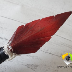 May include: Close-up of a spearhead with a dark red, feather-shaped blade. The blade is attached to a black leather-wrapped handle with white string. The spearhead is likely a decorative or cosplay item.