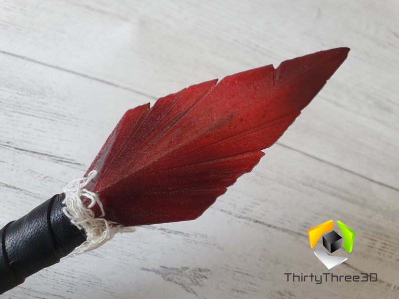 May include: Close-up of a spearhead with a dark red, feather-shaped blade. The blade is attached to a black leather-wrapped handle with white string. The spearhead is likely a decorative or cosplay item.