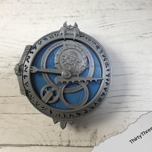 May include: A silver-coloured metal pendant with a blue centre and intricate details. The pendant features a circular design with a gear-like mechanism and runes around the edge.