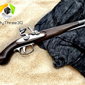 Puede incluir: Una r&eacute;plica de pistola de chispa con culata de madera marr&oacute;n oscuro y detalles met&aacute;licos plateados. El ca&ntilde;&oacute;n tiene una punta naranja. La pistola descansa sobre una superficie arenosa con un pa&ntilde;o gris oscuro.