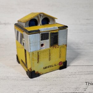 May include: A yellow and gray WALL-E robot toy with a red circle on the front. The robot has a small, rectangular body with a square head and two large, round eyes.