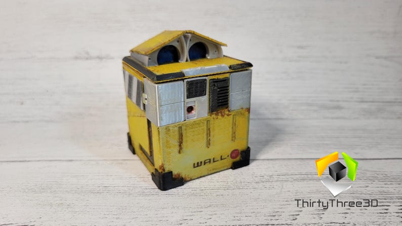 May include: A yellow and gray WALL-E robot toy with a red circle on the front. The robot has a small, rectangular body with a square head and two large, round eyes.