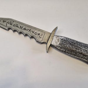 May include: A silver-colored dagger with a serrated edge and a wooden handle. The blade has etched markings. The handle is a dark gray and white color. The dagger is a prop or decorative item.