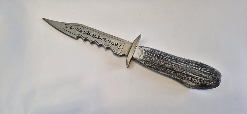 May include: A silver-colored dagger with a serrated edge and a wooden handle. The blade has etched markings. The handle is a dark gray and white color. The dagger is a prop or decorative item.