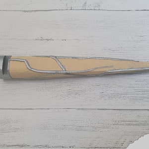 Op de afbeelding: Een beige en zilverkleurig dolk met een puntige punt en een getextureerd handvat. De dolk ligt op een wit houten oppervlak.