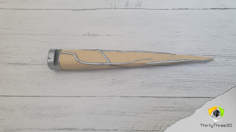 Op de afbeelding: Een beige en zilverkleurig dolk met een puntige punt en een getextureerd handvat. De dolk ligt op een wit houten oppervlak.