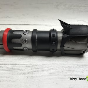 May include: A 3D-printed prop resembling a futuristic weapon. The device features a cylindrical body with a red ring, black accents, and metallic gray sections. It has a complex design with angular details and a fin-like structure on top.