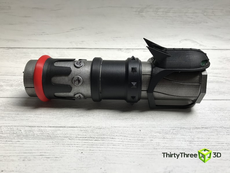 May include: A 3D-printed prop resembling a futuristic weapon. The device features a cylindrical body with a red ring, black accents, and metallic gray sections. It has a complex design with angular details and a fin-like structure on top.