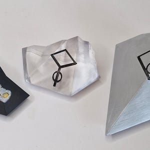 Pu&ograve; includere: Tre sculture geometriche astratte. Una &egrave; nera con dettagli oro e argento, un'altra &egrave; trasparente con un disegno nero e la terza &egrave; argentata con un disegno nero. Le sculture sono su una superficie bianca.