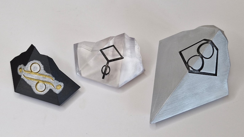 Pu&ograve; includere: Tre sculture geometriche astratte. Una &egrave; nera con dettagli oro e argento, un'altra &egrave; trasparente con un disegno nero e la terza &egrave; argentata con un disegno nero. Le sculture sono su una superficie bianca.