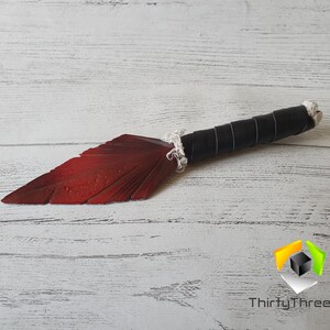 May include: A decorative knife with a red blade shaped like a feather. The handle is wrapped in black leather and white cord. The knife is lying on a white wooden surface.