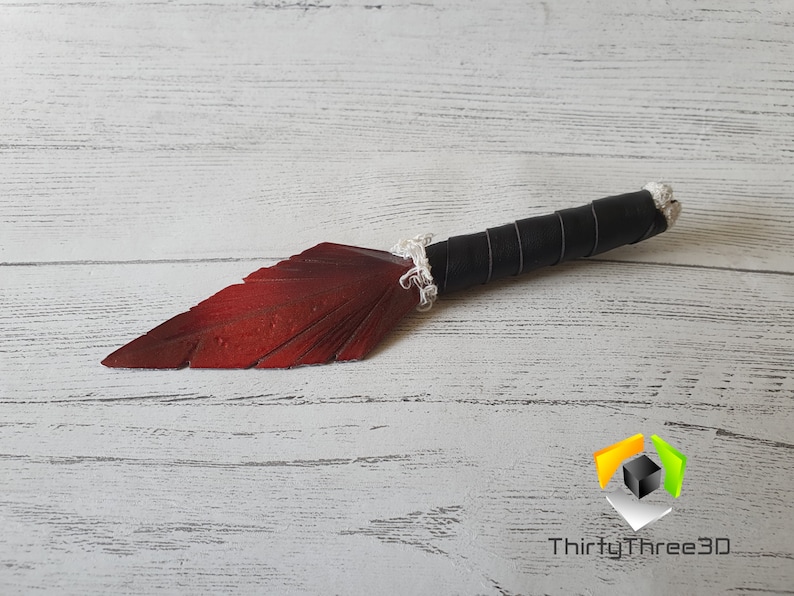 May include: A decorative knife with a red blade shaped like a feather. The handle is wrapped in black leather and white cord. The knife is lying on a white wooden surface.